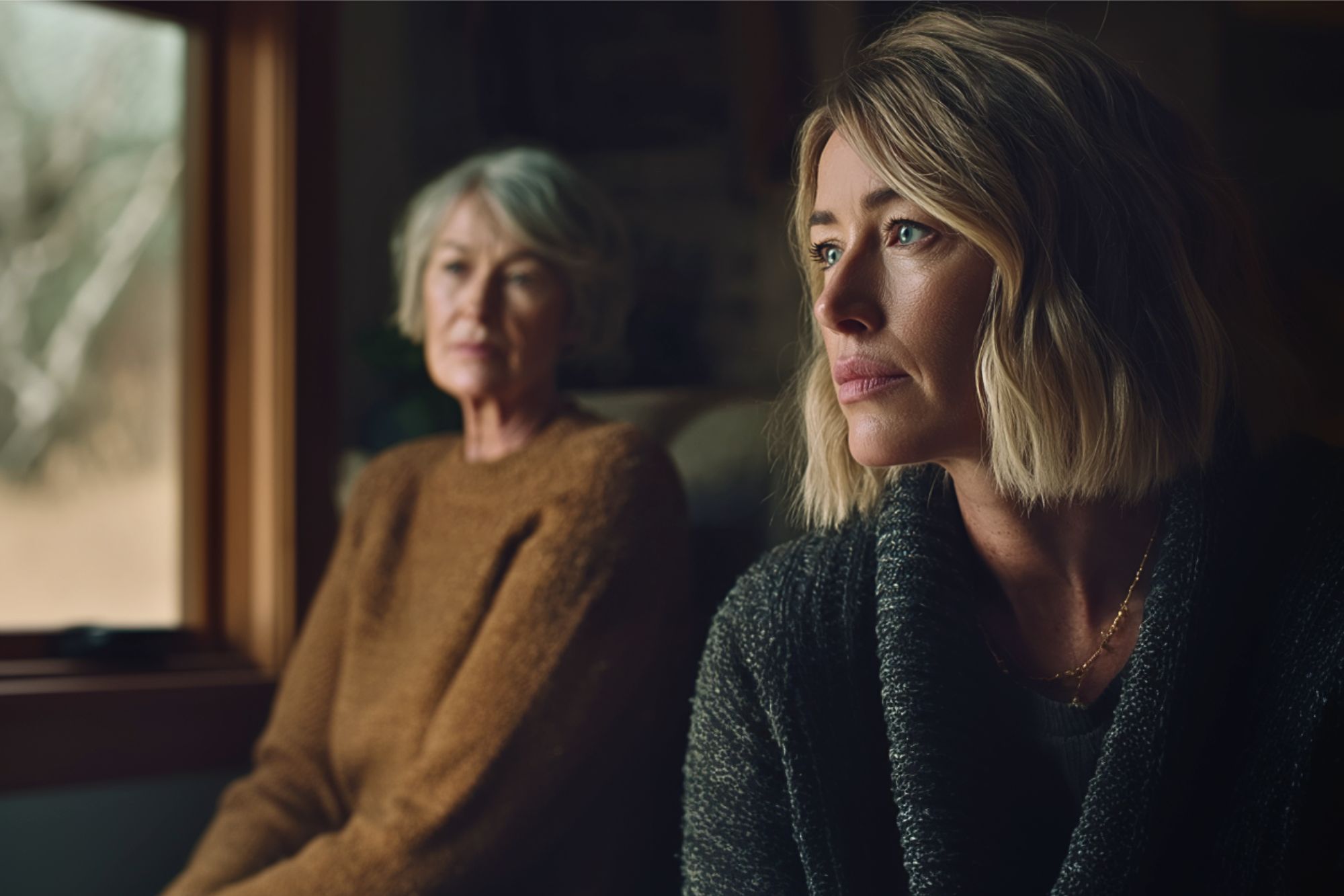 Two women sit indoors by a window. The woman in the foreground with blonde hair looks pensive, while an older woman with gray hair and a brown sweater sits in the background, slightly out of focus.