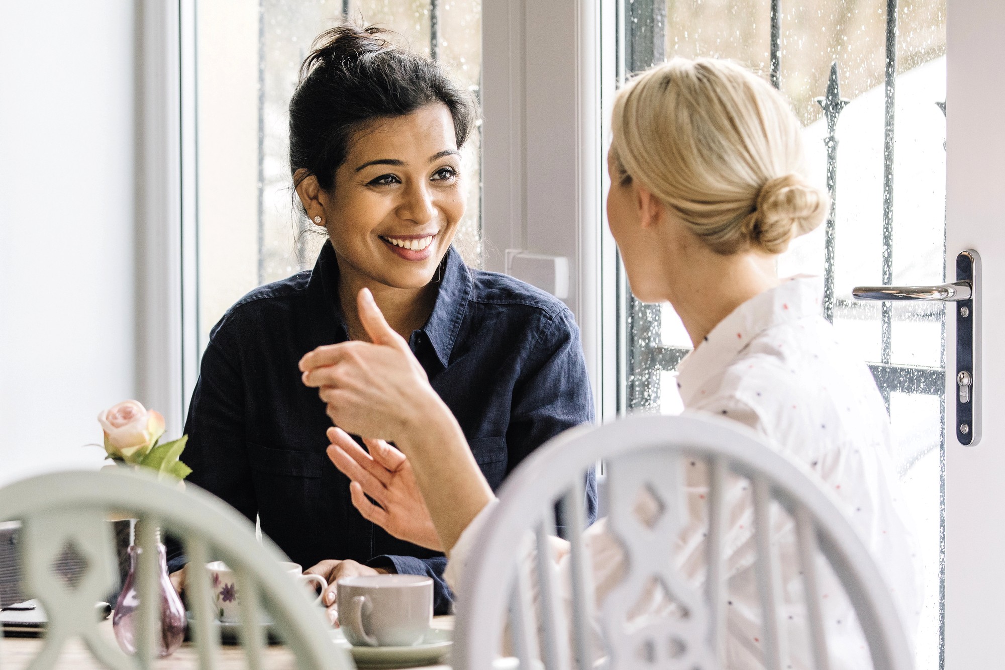 Two women sit at a table near a window, having a friendly conversation. One woman, with dark hair in a bun, smiles warmly at the other, who has blonde hair in a low bun. A cup and a vase with a rose are on the table.