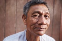 A middle-aged man with short gray hair and a slight smile looks at the camera, standing in front of a wooden background and wearing a light-colored shirt.