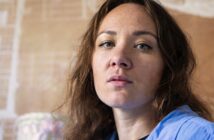 A woman with wavy brown hair and light eyes looks directly at the camera with a neutral expression, wearing a blue top. The background is softly blurred in warm tones.