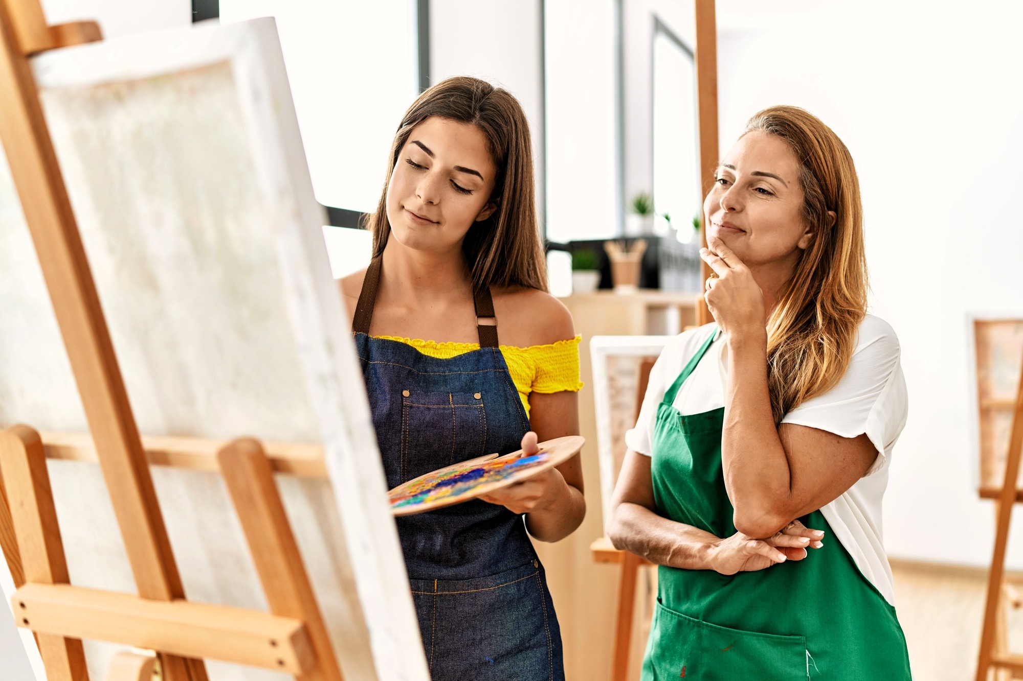 Two women stand by an easel in a bright art studio. One holds a palette and brush while painting, and the other, wearing an apron, watches thoughtfully with her hand on her chin.