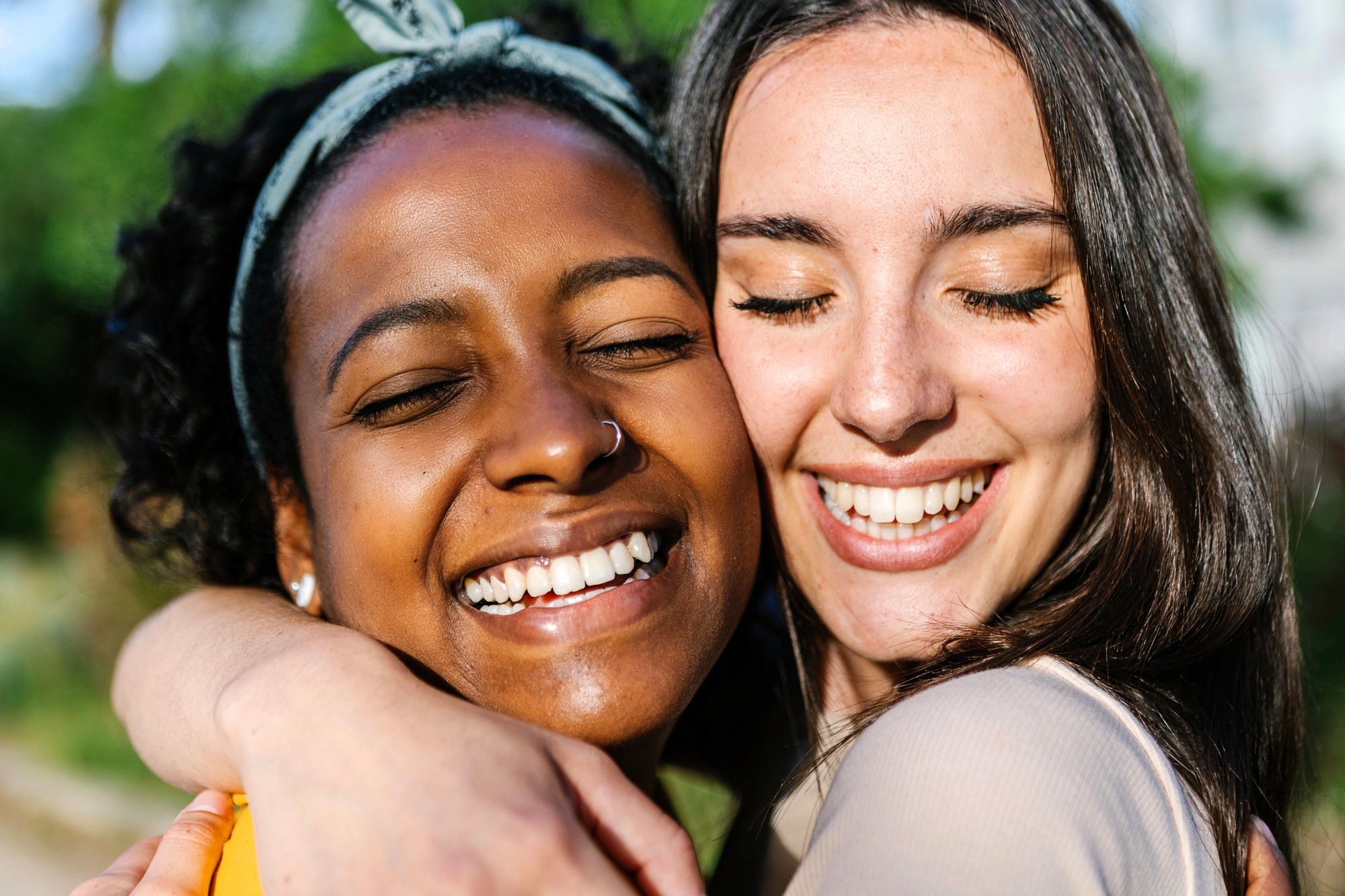 Two young women smiling with eyes closed, embracing each other outdoors on a sunny day, showing joy and friendship.