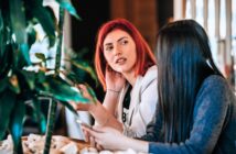 Two women are sitting indoors at a table, engaged in conversation. One has red hair and gestures while speaking; the other has long dark hair and holds a smartphone. Green plants are visible in the foreground.