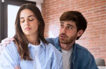 A man tries to comfort a woman who looks upset and turns away from him, crossing her arms. They are indoors with a brick wall in the background.
