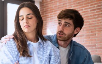A man tries to comfort a woman who looks upset and turns away from him, crossing her arms. They are indoors with a brick wall in the background.