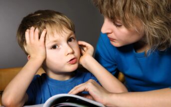 A young boy looks frustrated while sitting at a table with an open book. An adult beside him, wearing a blue shirt, offers support, gently touching his arm and looking at him with concern.