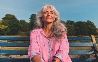 A smiling older woman with long gray hair sits on a blue bench outdoors, wearing a pink blouse with floral embroidery, with trees and a clear sky in the background.