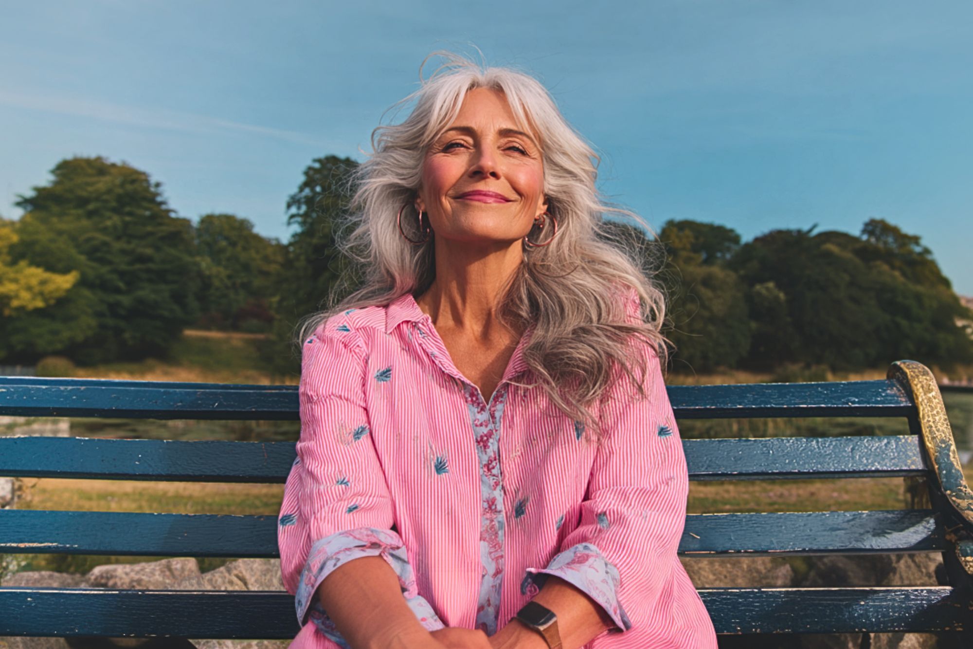 A smiling older woman with long gray hair sits on a blue bench outdoors, wearing a pink blouse with floral embroidery, with trees and a clear sky in the background.