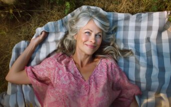 A smiling woman with gray hair in a pink patterned blouse lies on a checkered blanket outdoors, surrounded by dry grass and greenery, enjoying a peaceful moment.