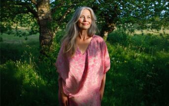 A woman with long gray hair in a pink dress stands in a sunlit, green outdoor setting with trees and grass, smiling and looking to the side.