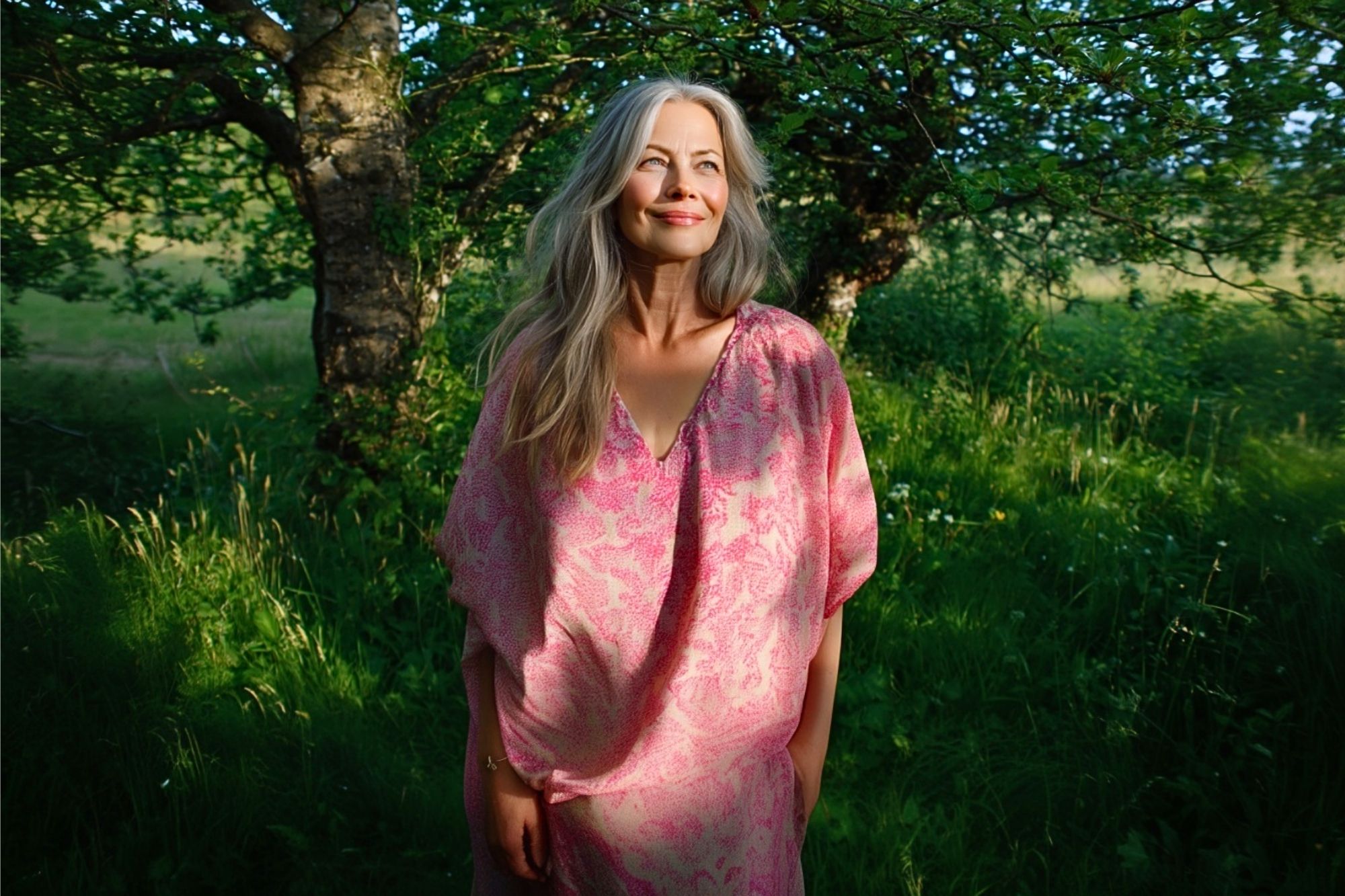 A woman with long gray hair in a pink dress stands in a sunlit, green outdoor setting with trees and grass, smiling and looking to the side.
