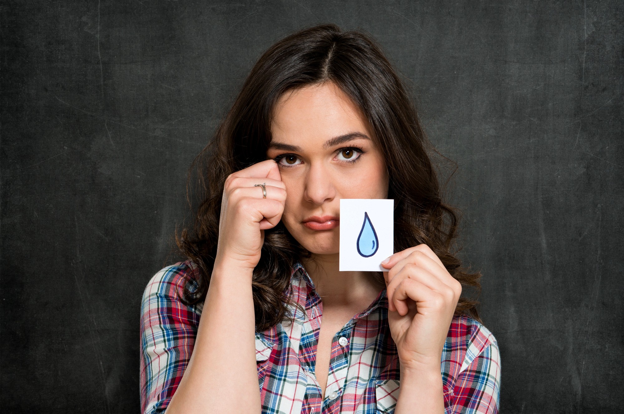 A young woman in a plaid shirt stands in front of a chalkboard, holding a paper with a blue teardrop drawn on it. She makes a sad face and gestures as if wiping a tear from her eye.