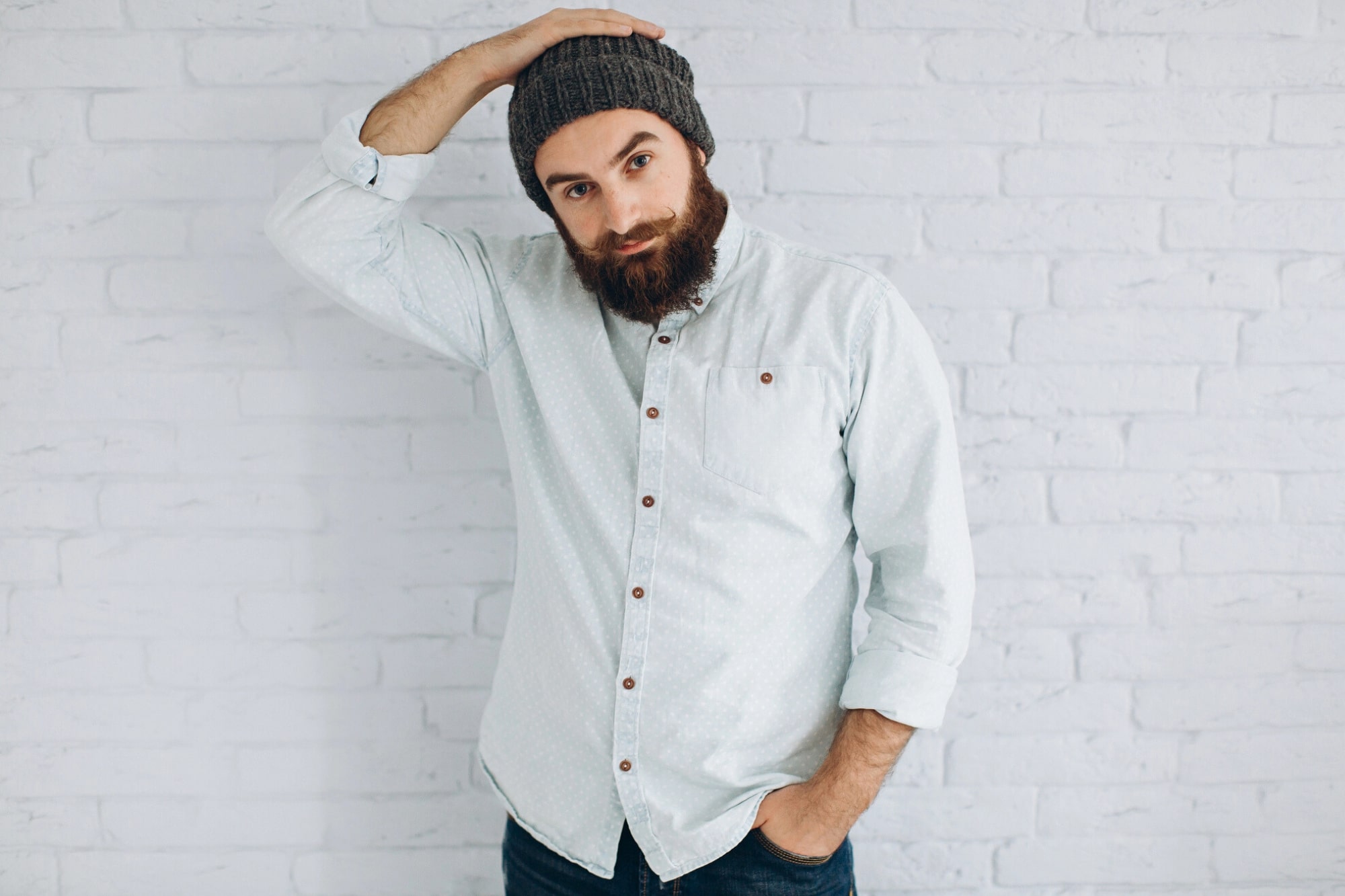 A bearded man in a gray knit beanie and light button-up shirt stands against a white brick wall, looking at the camera while holding one hand on top of his head.