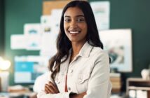 A woman with long dark hair and a light-colored jacket smiles confidently with her arms crossed, standing in an office with a dark green wall and blurred bulletin board in the background.