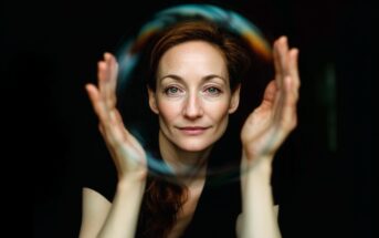 A woman with auburn hair gazes forward, holding her hands up around a large, translucent soap bubble against a dark background. The bubble appears to frame her face.