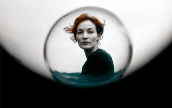 A woman with red hair is seen through a glass sphere, creating a circular frame around her face. The background is muted, highlighting her contemplative expression.