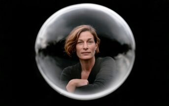 A woman looking forward, framed within a circular glass sphere against a black background.