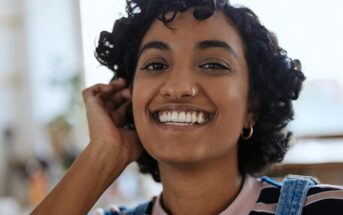 A young woman with short curly hair and hoop earrings smiles warmly at the camera, touching her hair. She is wearing a striped shirt and denim overalls. The background is softly blurred.