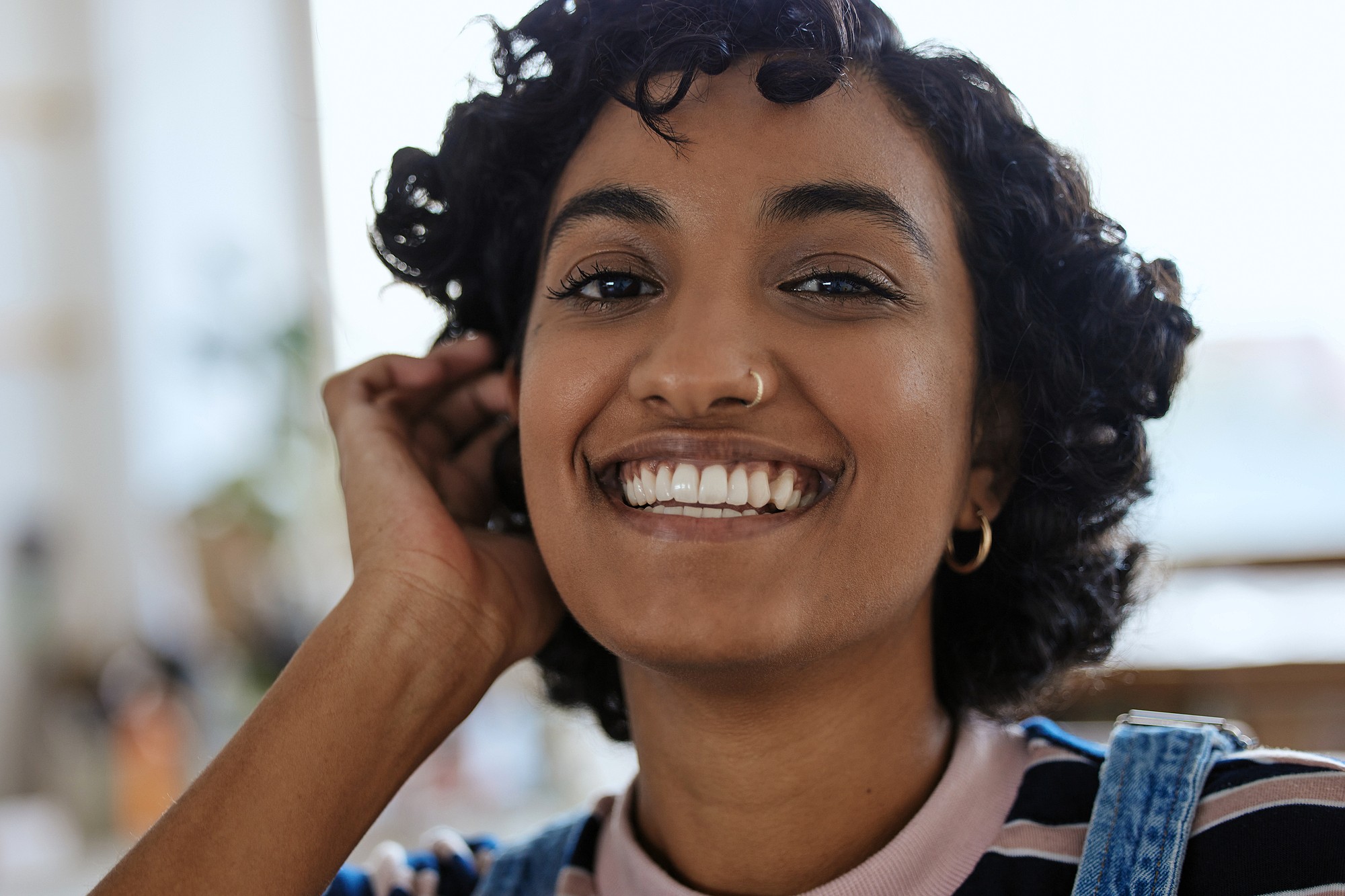 A young woman with short curly hair and hoop earrings smiles warmly at the camera, touching her hair. She is wearing a striped shirt and denim overalls. The background is softly blurred.