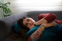A woman with long brown hair and a tattooed arm lies on a gray couch, hugging a teal pillow and looking thoughtfully ahead. Soft daylight filters in through a window behind her.