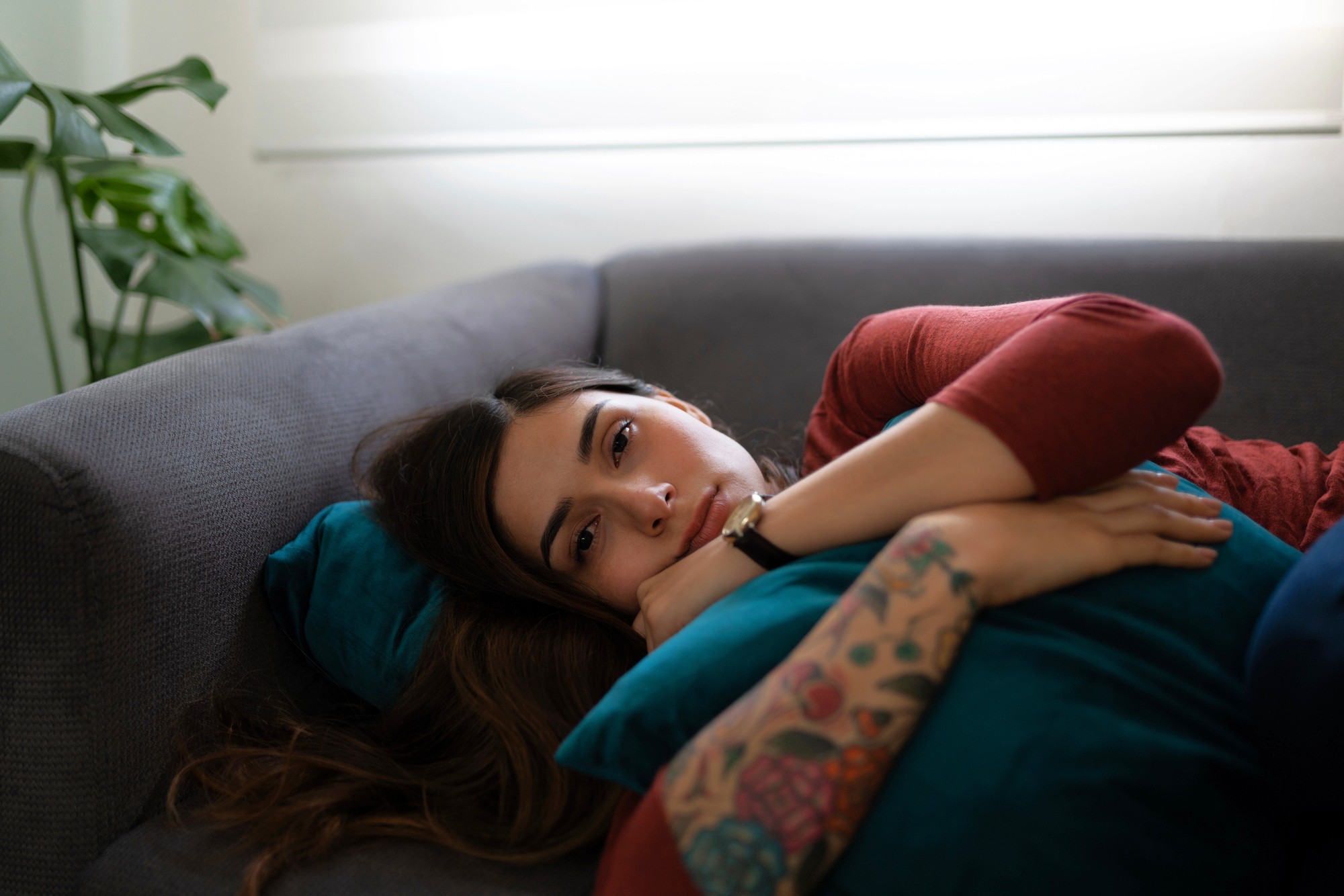 A woman with long brown hair and a tattooed arm lies on a gray couch, hugging a teal pillow and looking thoughtfully ahead. Soft daylight filters in through a window behind her.