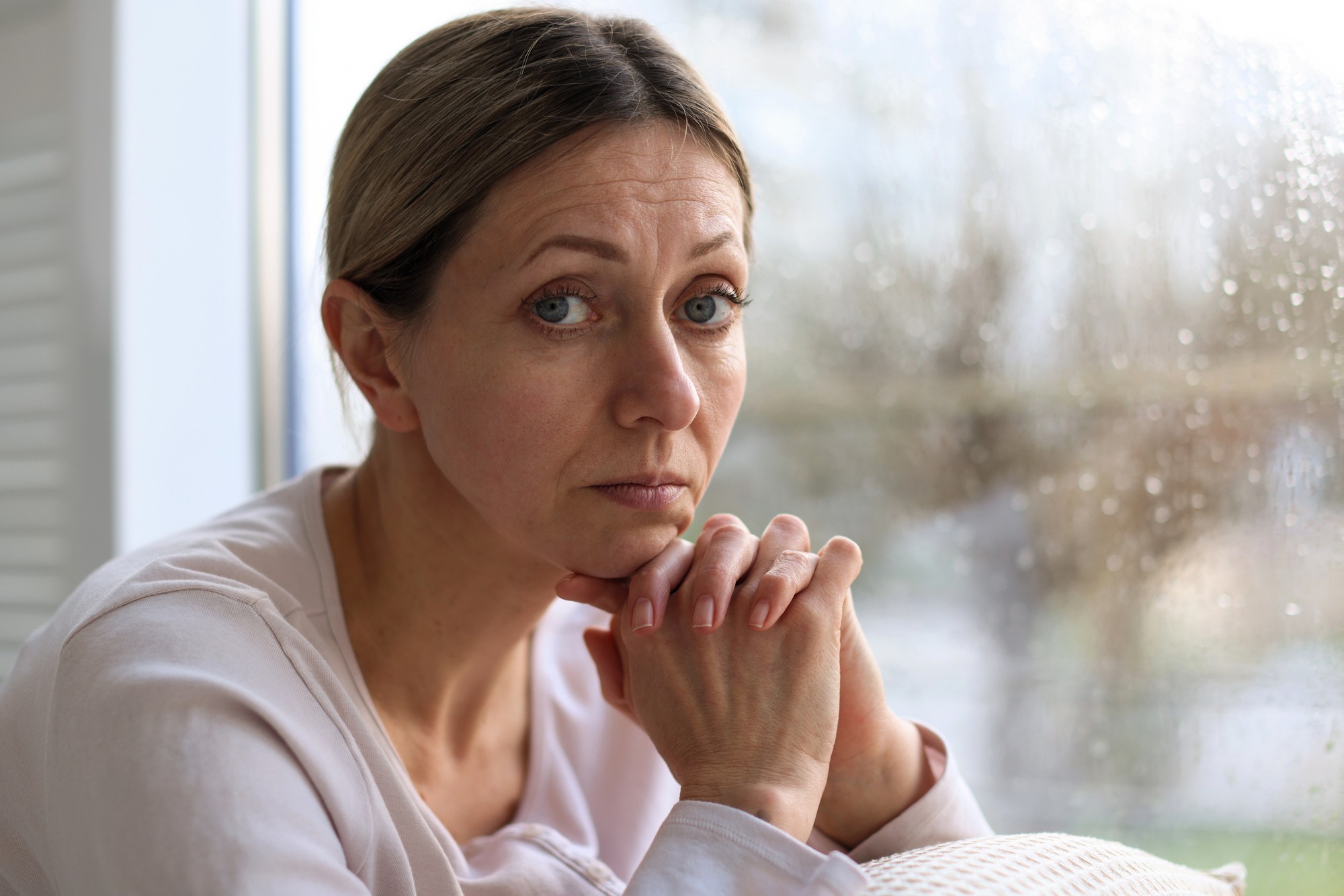 A woman with light brown hair pulled back rests her chin on her hands and looks thoughtfully at the camera. She sits by a window with raindrops visible on the glass, creating a somber atmosphere.