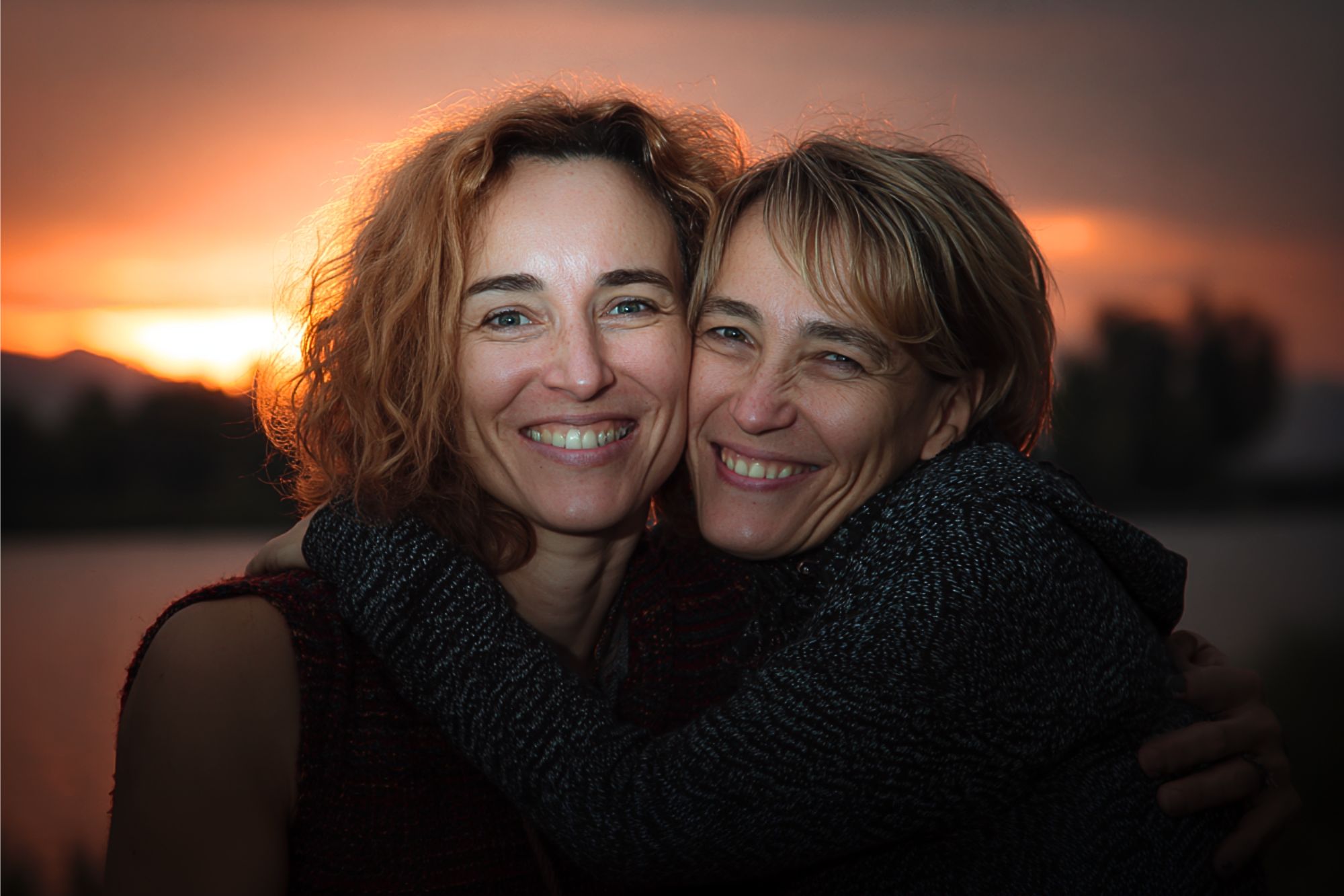 Two women smile warmly and embrace each other outdoors at sunset, with a glowing sky and blurred trees and water in the background.