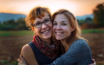 Two women smiling and hugging outdoors at sunset, with a blurred landscape and mountains in the background.