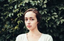 A young woman with light skin and brown hair stands in front of a wall covered in green leafy plants, looking directly at the camera with a neutral expression. She is wearing a white top.
