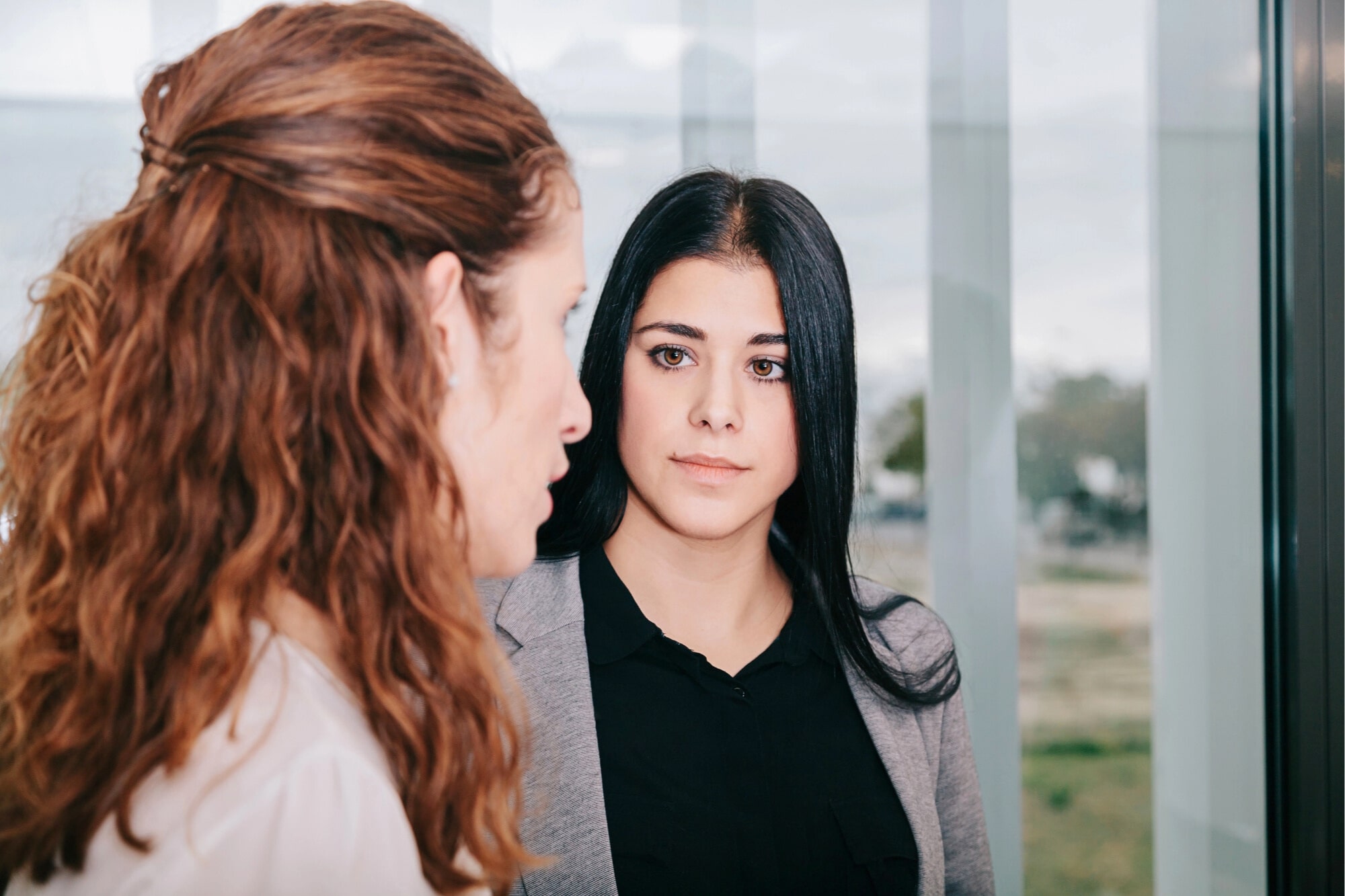 Two women are having a serious conversation indoors. One woman with long brown hair is facing away from the camera, while the other woman with straight black hair and a gray blazer looks at her attentively.