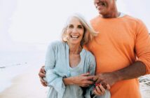 An older woman with long gray hair smiles while standing on a beach, holding hands with a man wearing an orange shirt. The sea and sand are visible in the background.