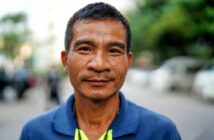 A middle-aged man with short hair and a slight smile stands outdoors, wearing a blue collared shirt. The background is blurred, showing trees and cars.