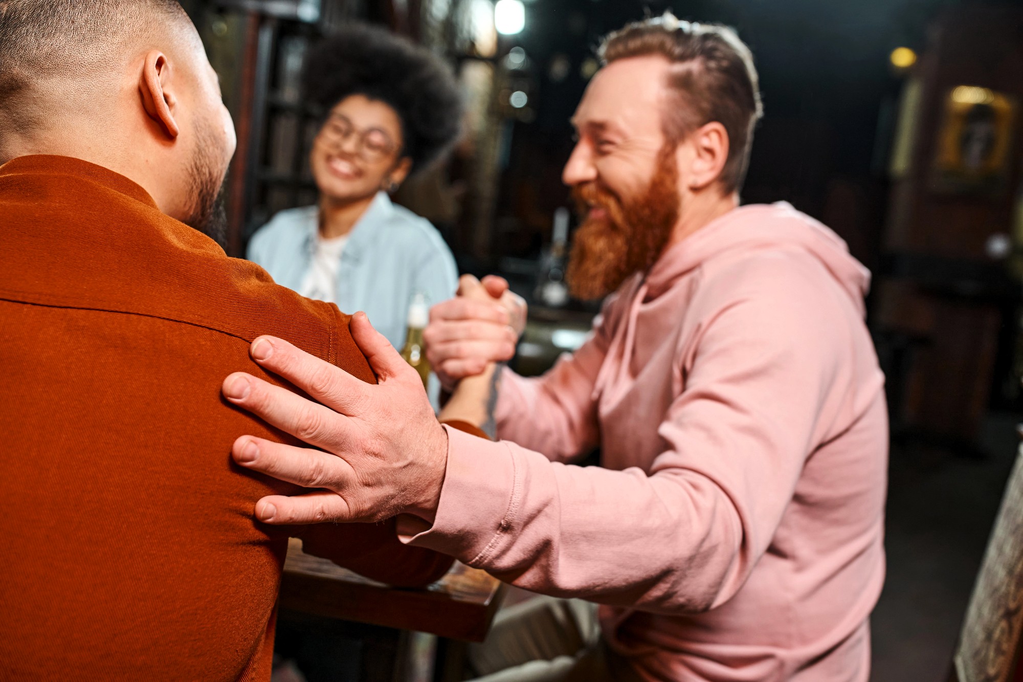 Two people are arm wrestling at a table while smiling, and a third person in the background is watching and smiling. The scene appears friendly and relaxed in a dimly lit setting.