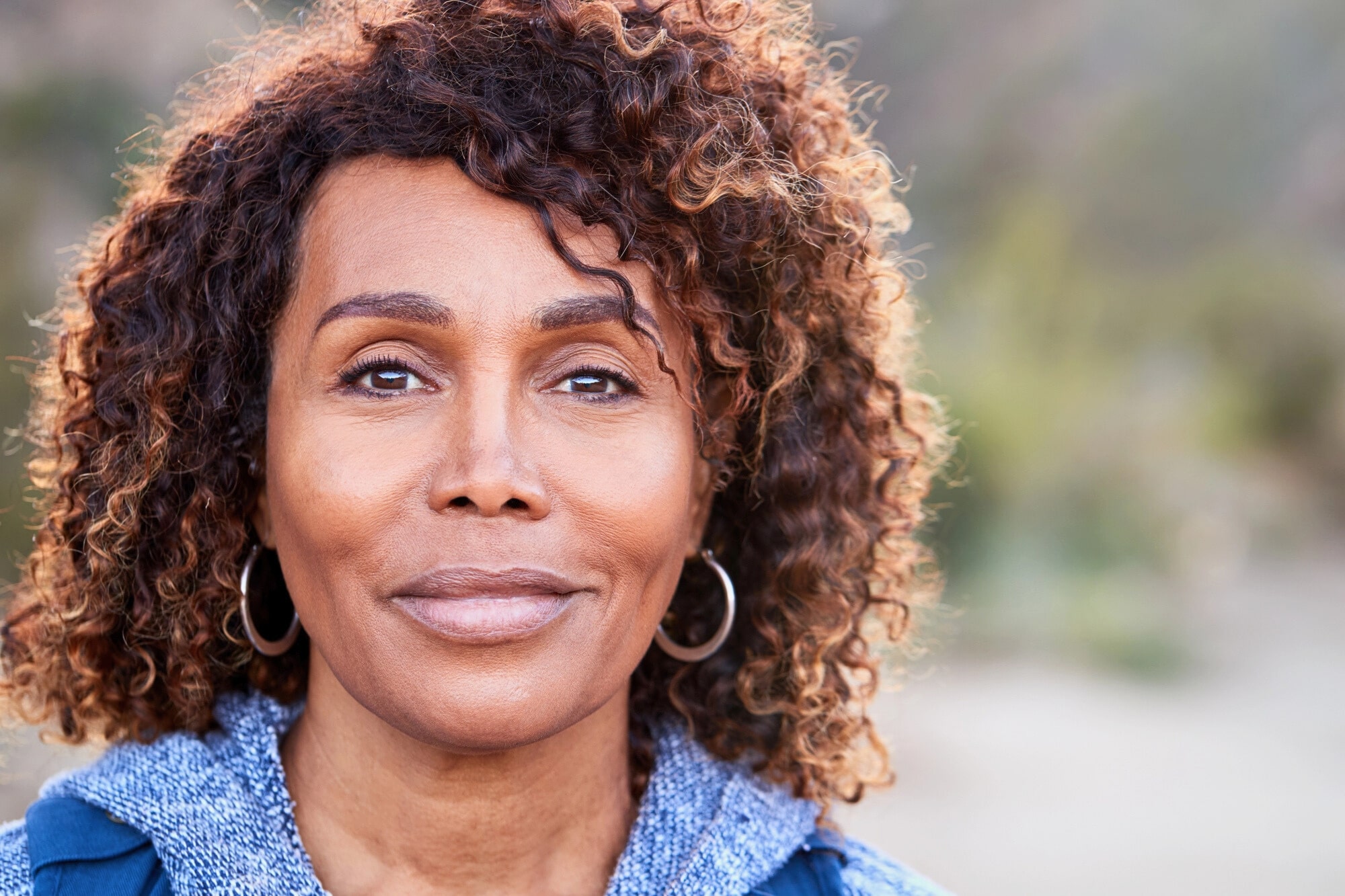 A middle-aged woman with curly hair and hoop earrings looks calmly at the camera outdoors, wearing a blue sweater. The background is softly blurred with natural greenery.