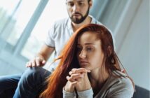 A woman with long red hair sits looking sad and thoughtful, with her hands clasped. A man sits behind her, gently touching her shoulder, appearing concerned and supportive.