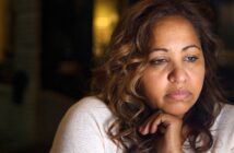 A woman with wavy brown hair rests her chin on her hand, looking thoughtful and sad. She is wearing a light-colored sweater, with a soft, dim background behind her.