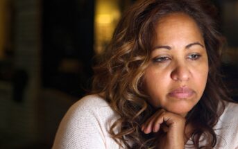 A woman with wavy brown hair rests her chin on her hand, looking thoughtful and sad. She is wearing a light-colored sweater, with a soft, dim background behind her.