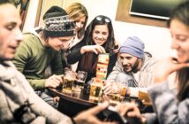 Five friends sit around a table with drinks, smiling and playing a game of Jenga. One woman is carefully removing a block while the others watch and laugh. The group appears relaxed and is warmly dressed.