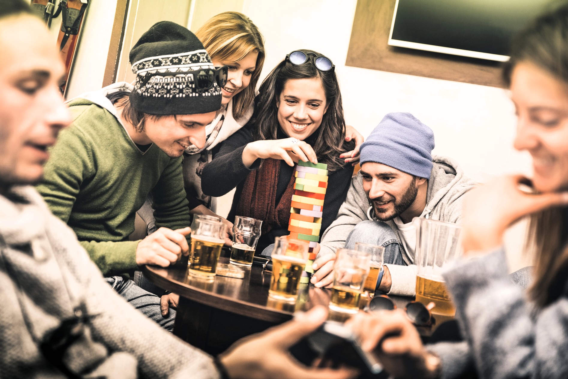 Five friends sit around a table with drinks, smiling and playing a game of Jenga. One woman is carefully removing a block while the others watch and laugh. The group appears relaxed and is warmly dressed.