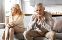An older man and woman sit on a couch, both looking upset and facing away from each other. The man looks down with his hands clasped under his chin, while the woman rests her face on her hand, staring into the distance.