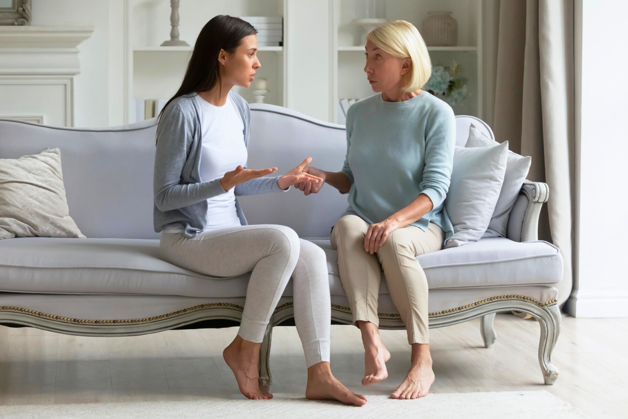 Two women sit on a sofa in a living room, facing each other and having an intense conversation. Both have serious expressions and use hand gestures, suggesting a disagreement or emotional discussion.