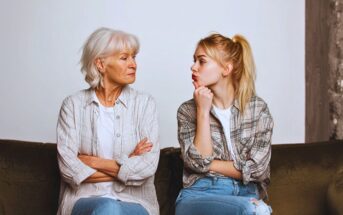 An older woman and a young woman sit on a couch with arms crossed, facing each other. The young woman makes a pouty face, while the older woman looks at her seriously. Both wear casual clothes and similar patterned shirts.