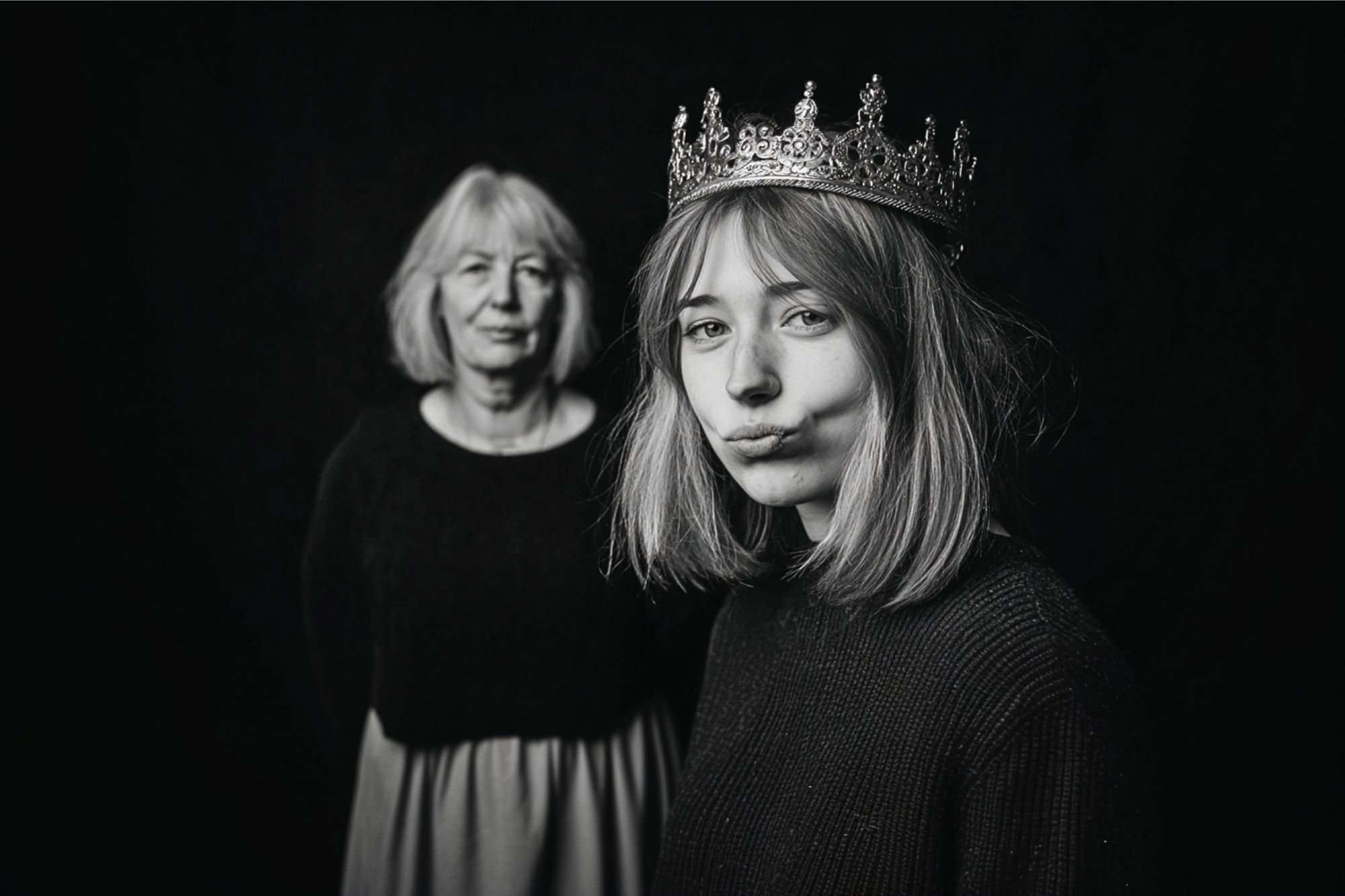 A young woman wearing a crown stands in the foreground making a playful expression, while an older woman stands behind her, both against a dark background. The photo is in black and white.