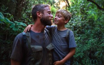 A bearded man and a young boy, both wearing backpacks and casual clothes, stand close together in a lush, green forest, smiling warmly at each other as sunlight filters through the trees.