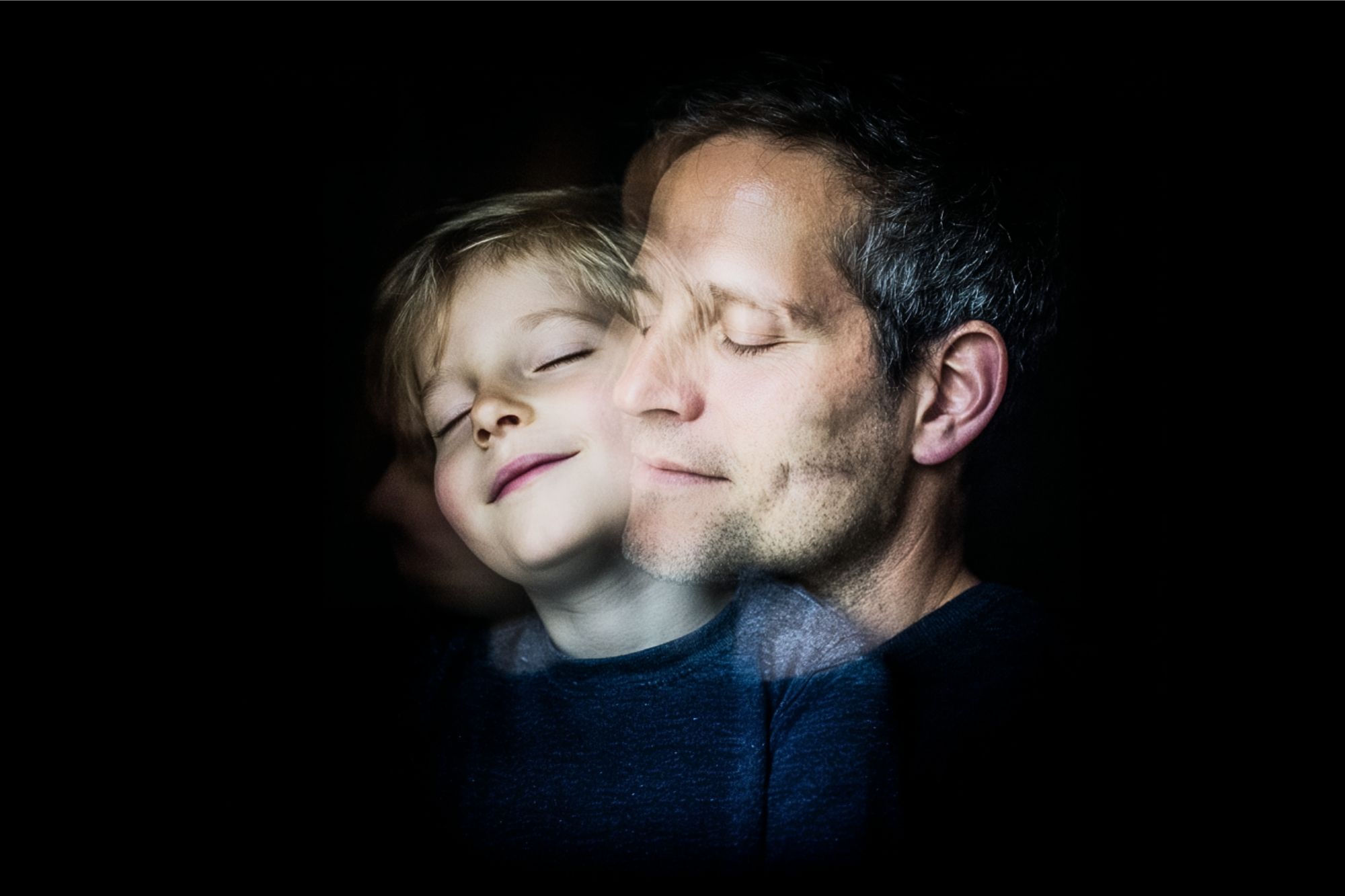 A child and an adult, both with closed eyes and peaceful expressions, overlap in a double exposure effect against a dark background.