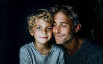 A smiling man with closed eyes hugs a young boy with blue eyes and curly blond hair. Both wear gray shirts and are lit softly against a dark background.