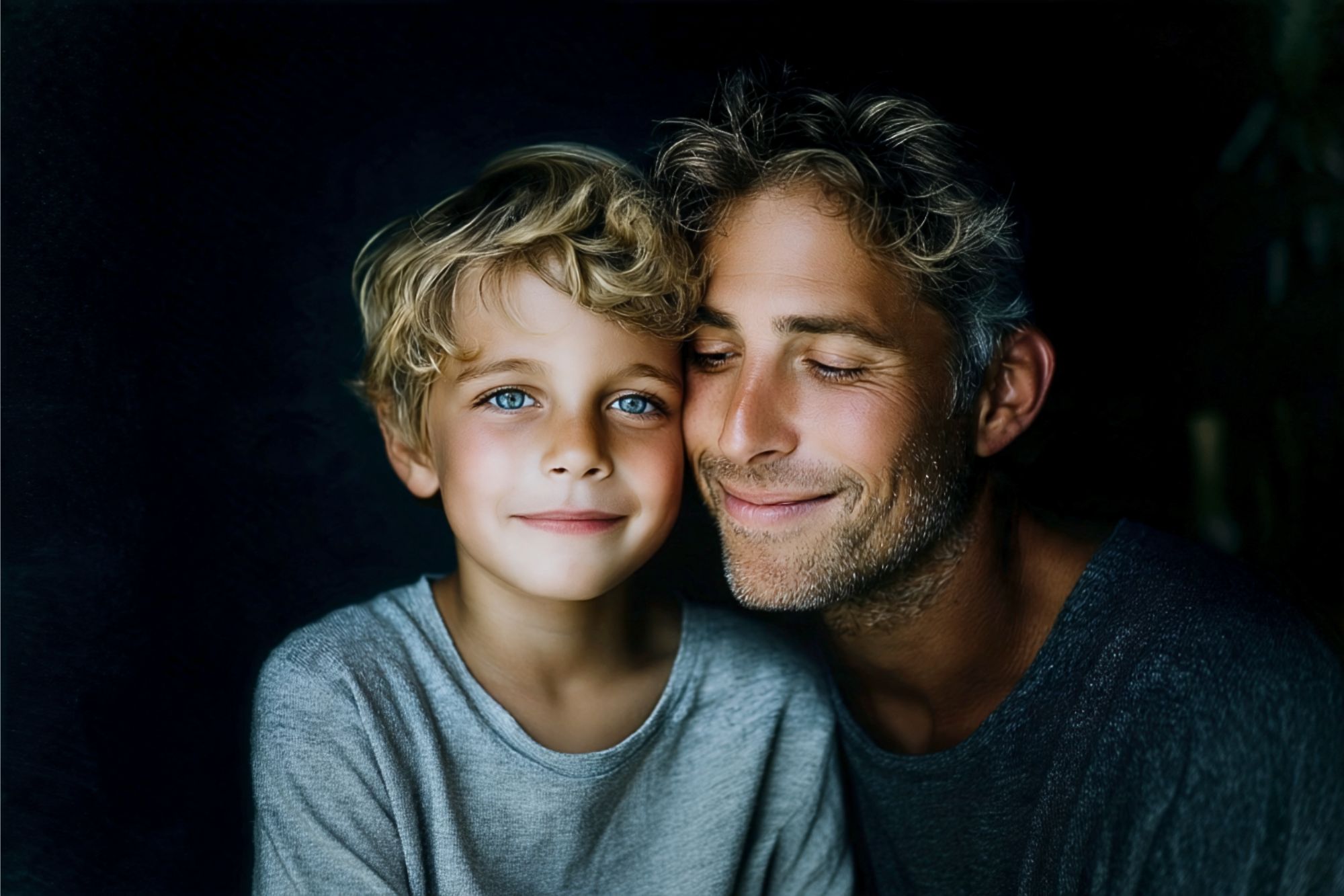 A smiling man with closed eyes hugs a young boy with blue eyes and curly blond hair. Both wear gray shirts and are lit softly against a dark background.