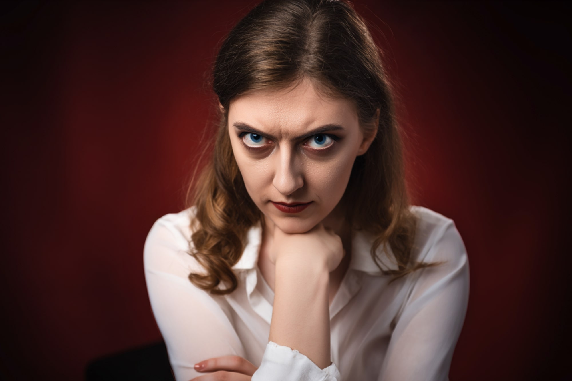 A woman with long brown hair and a white blouse sits with her chin resting on her fist, staring intensely and frowning with a serious, angry expression against a dark red background.