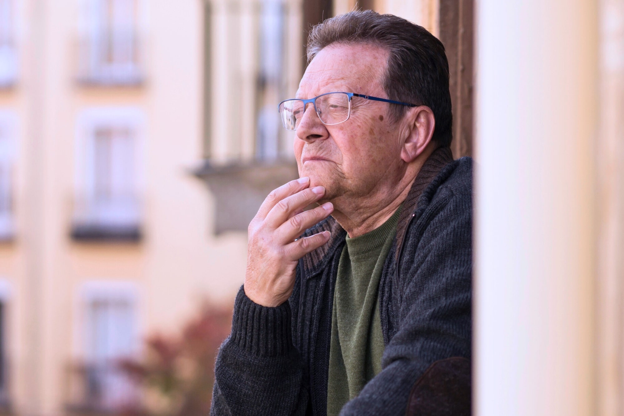 An older man with glasses and a thoughtful expression rests his hand on his chin while looking out from a balcony. He wears a dark sweater and stands against a blurred background of building windows.
