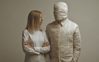 A woman in a white shirt stands beside a person wrapped in newspaper from head to toe, including their face, with only their hands exposed. Both face forward, the woman looking at the person in newspaper.